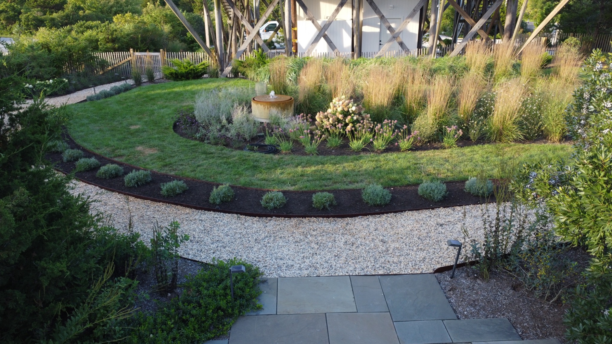 A bubbling garden fountain among flowering plants and tall grasses, framed by lawn and a curved gravel path.